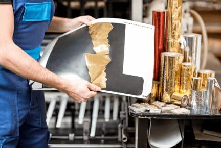 A man in overalls stands beside a hot foil printing machine, holding a piece of paper in his hands.